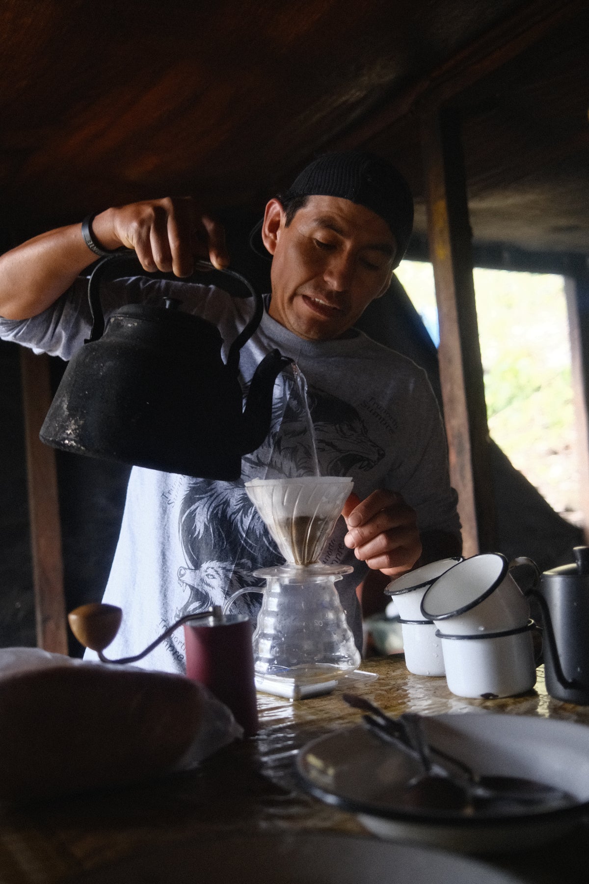 A man in a rustic kitchen pouring hot water from a black kettle into a glass pour-over coffee dripper. White ceramic mugs and a coffee grinder are visible on the table in a dimly lit, wood-paneled room.