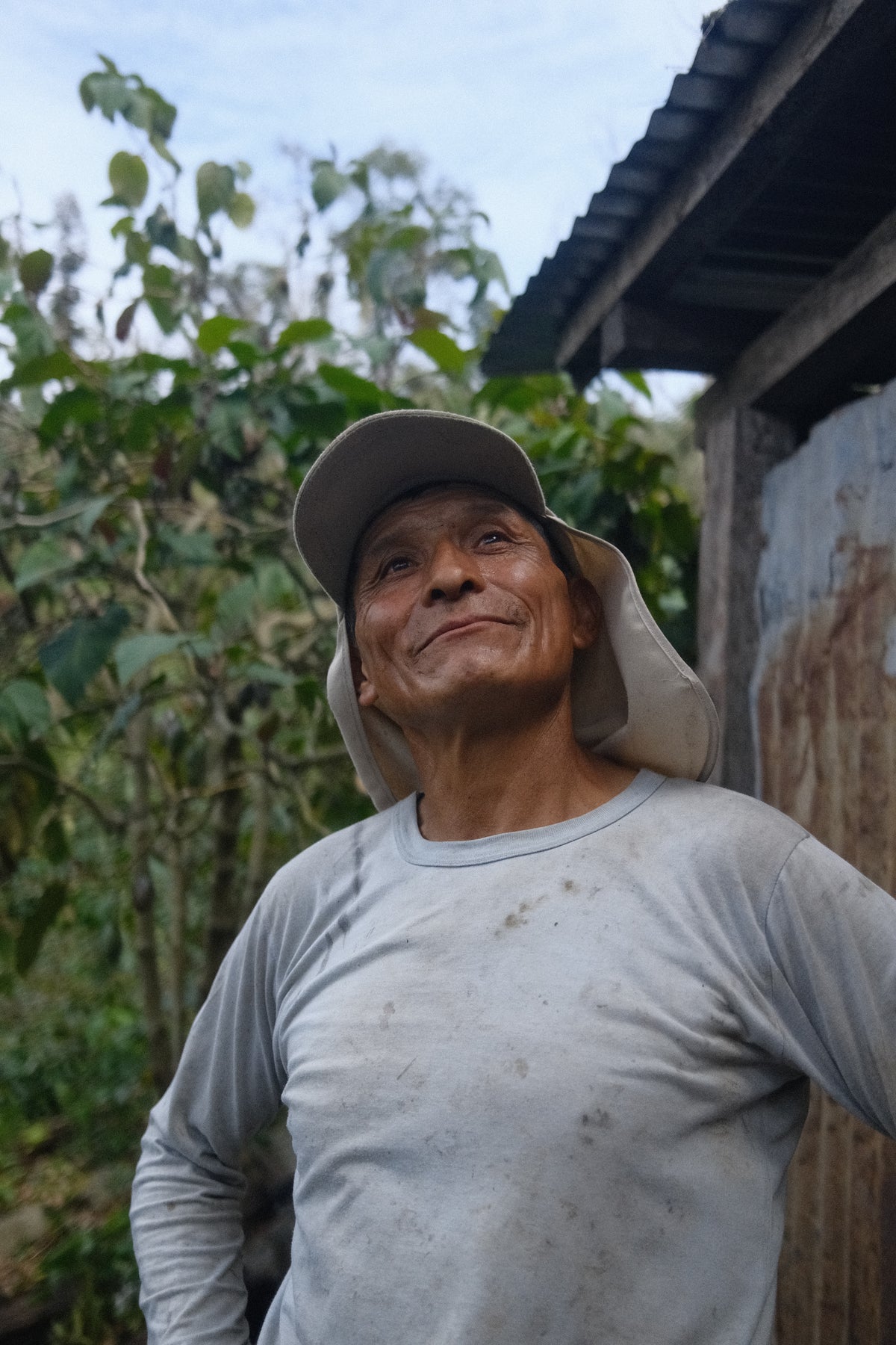A smiling man wearing a light gray long-sleeve shirt and a beige sun hat with a neck flap, standing outdoors next to a rustic corrugated metal wall and green foliage.